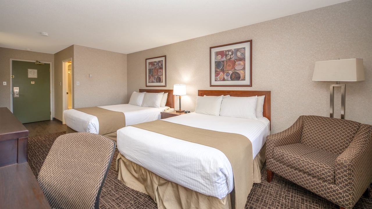 Two beds, chairs, nightstands with lamps, and framed artwork on walls in a hotel room.