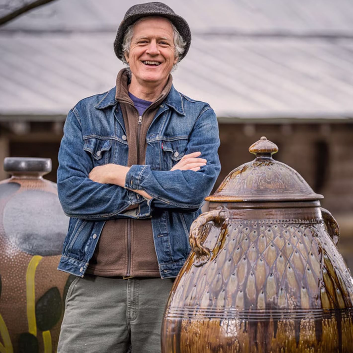Smiling potter in denim jacket and hat standing beside large decorative ceramic vessel outdoors at pottery studio.