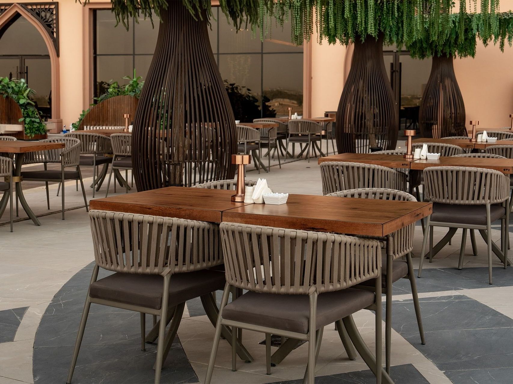 Outdoor dining area with wooden tables and chairs under artificial trees at World Heart Hotel Baghdad.