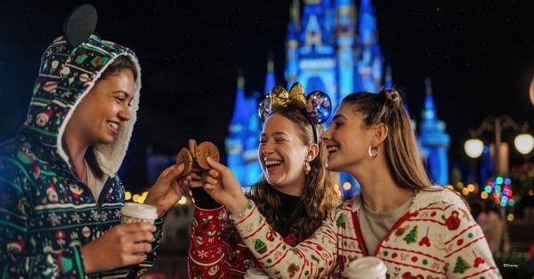 Three young women in cozy holiday attire and Minnie ears toast with cookies and cocoa in front of Cinderella Castle during Mickey's Very Merry Christmas Party. 