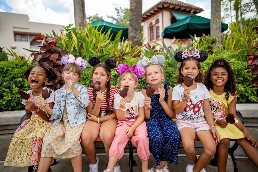 Seven girls wearing Minnie ears in a row eating Mickey ice cream bars outdoors. 