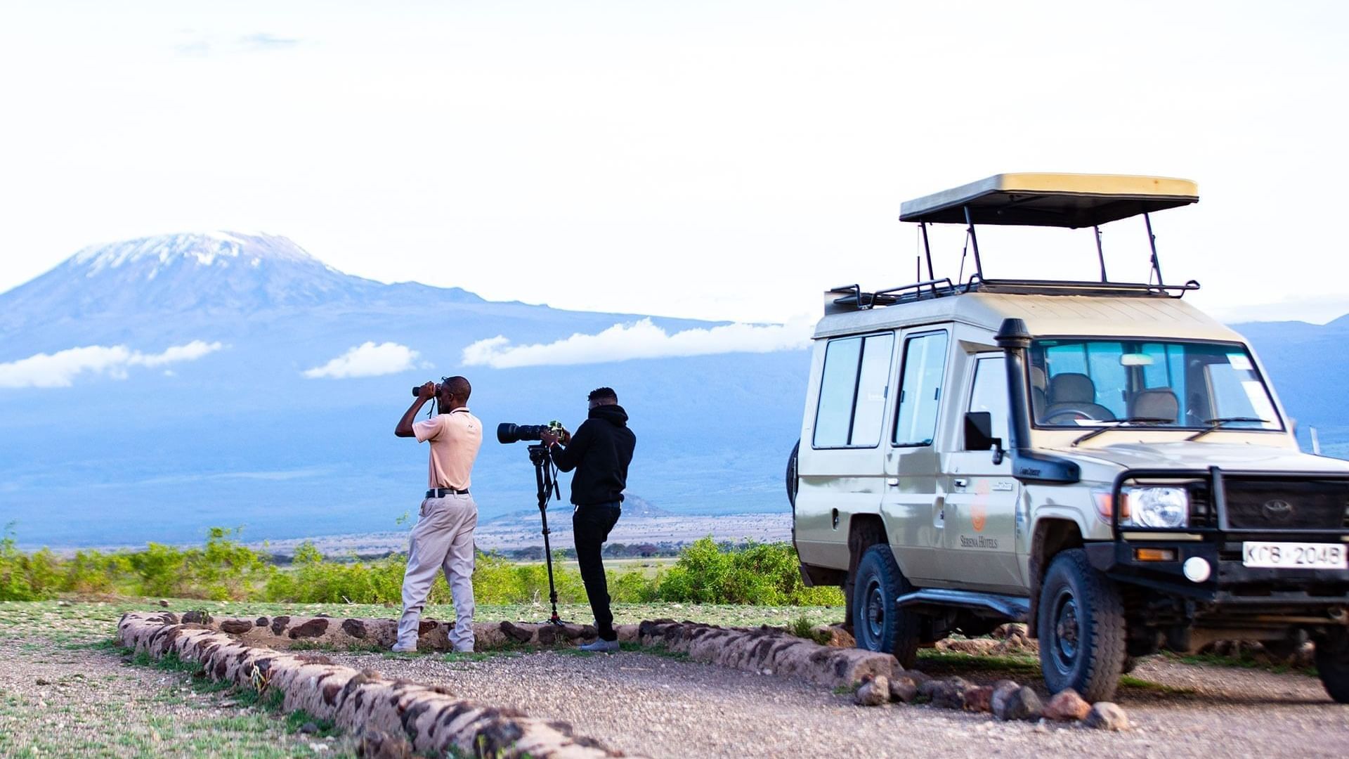 Two guides with binoculars and a camera on a tripod standing by a vehicle facing Mt Kilimanjaro at Serena Hotels & Resorts