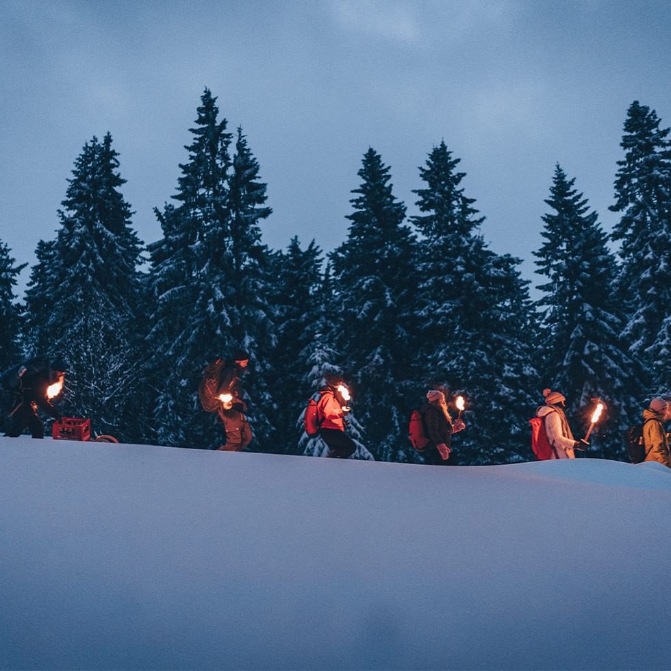Group of hikers with torches in a snowy forest at dusk for a torchlight hike.