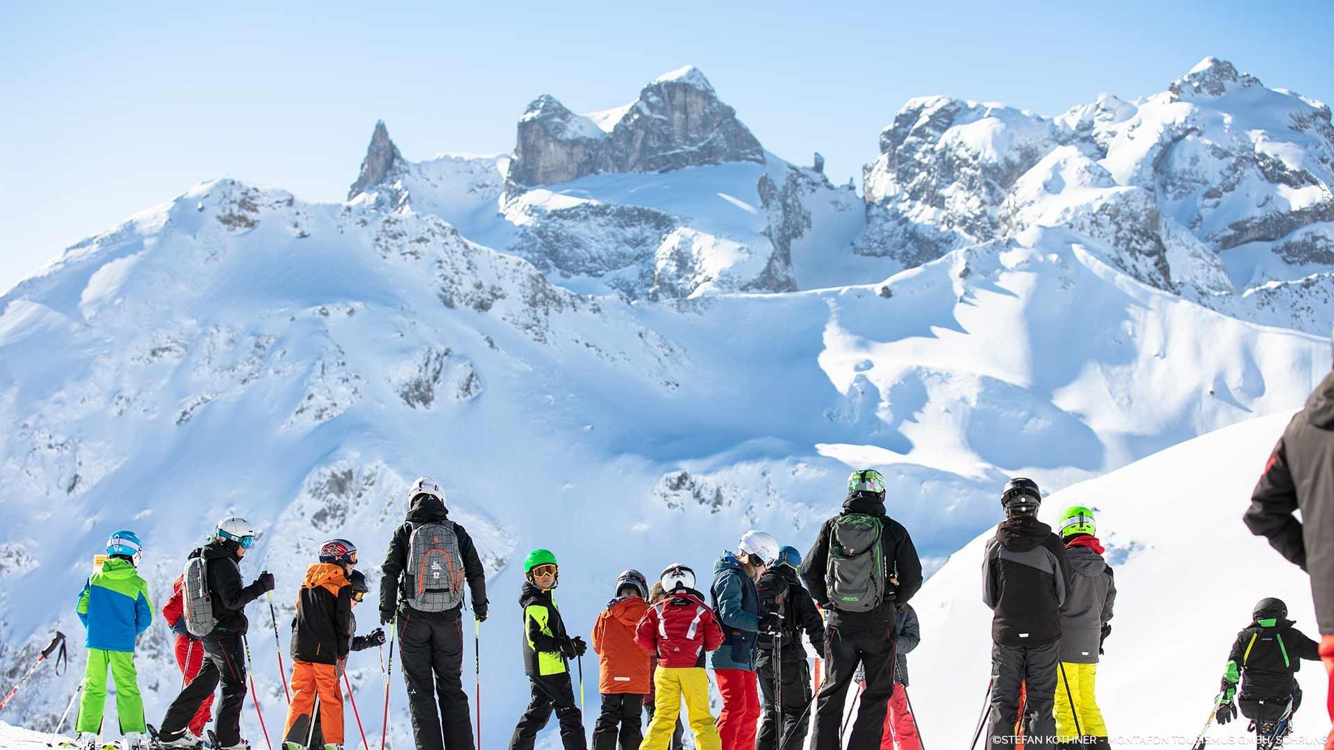 Gruppo di sciatori con attrezzatura in una stazione sciistica alpina con montagne innevate sullo sfondo.