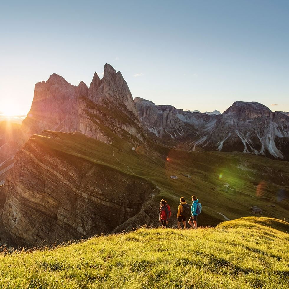 Drei Wanderer mit Rucksäcken auf einer Bergwiese vor einer Gebirgskette bei Sonnenuntergang.