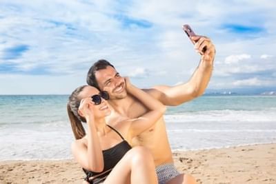 Smiling couple taking a selfie on a sandy beach with ocean and cloudy sky at Playa Los Arcos Puerto Vallarta
