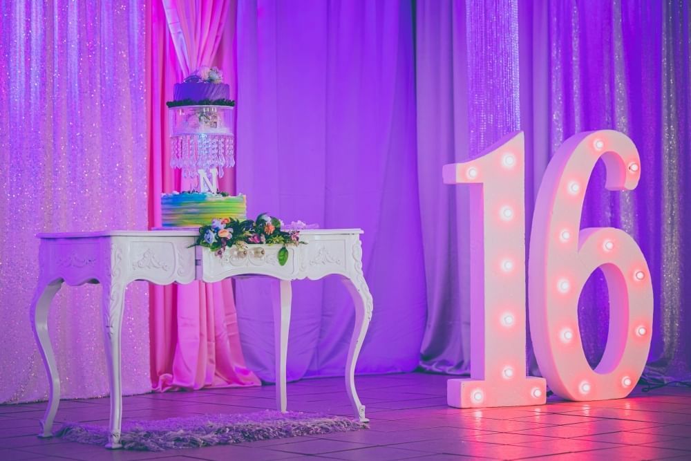 A white vanity table with a tiered cake stands beside a large number 16 with party lights, both in front of hanging drapery lit pink and purple. 