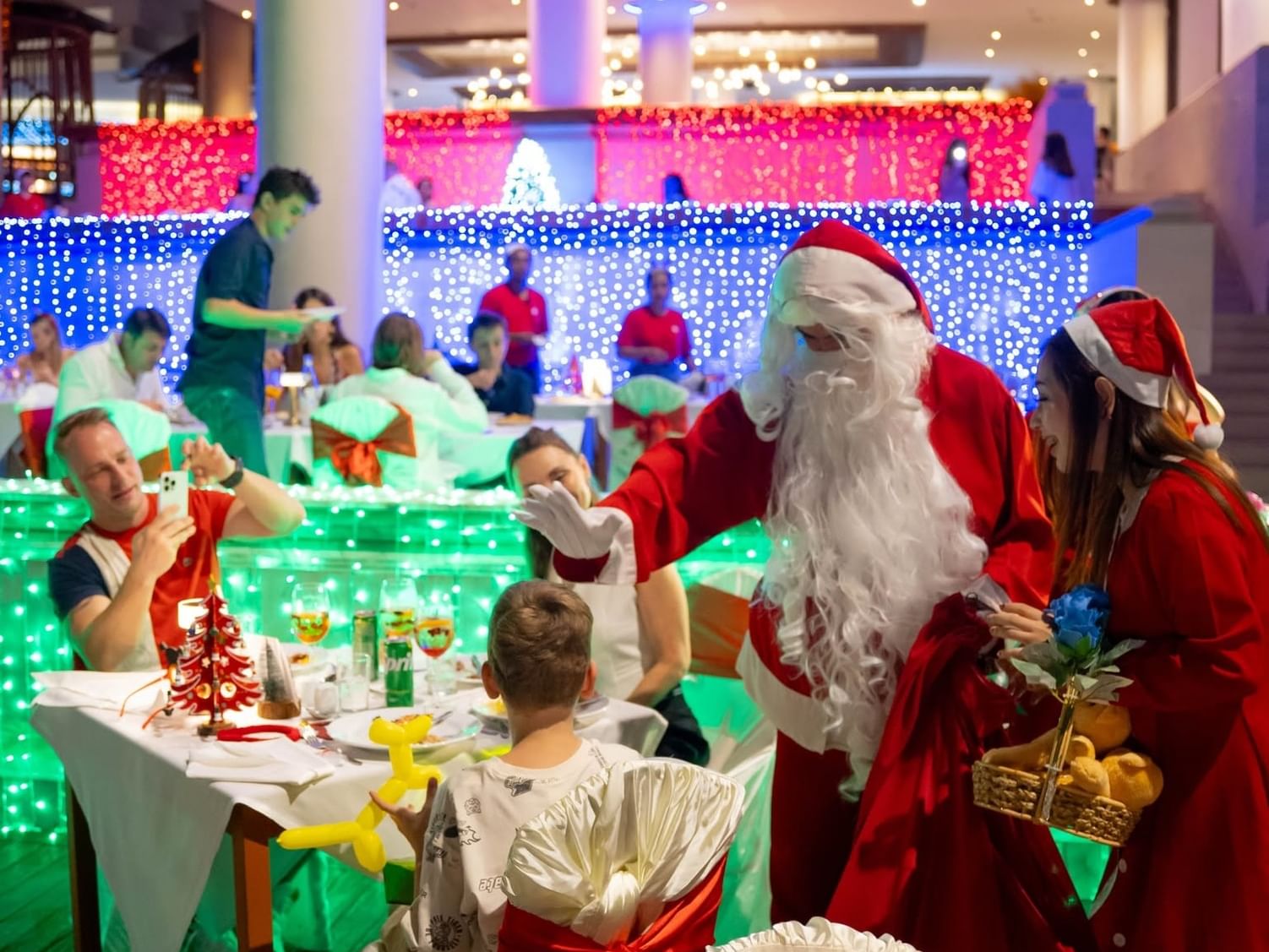 Santa Claus greeting families at festive dining tables with colorful lights and holiday decorations in the background.