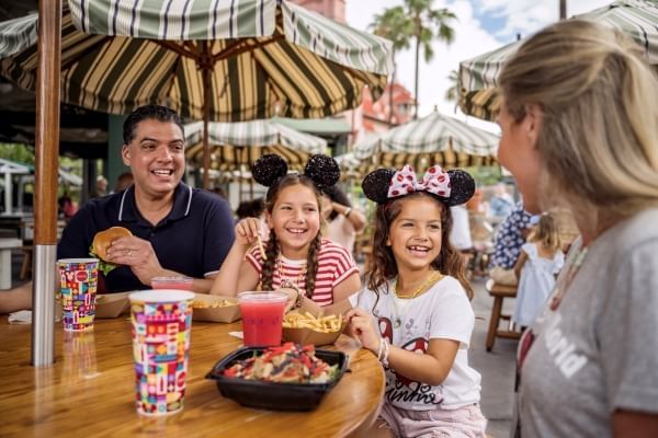 Two adults and two girls in Minnie ears eat at a table outdoors beneath striped umbrellas. 