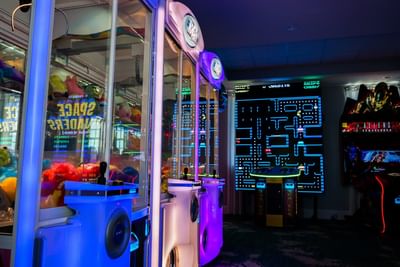 Neon-lit arcade room with crane machines, Pac-Man light art, and classic video games for families at Centennial Plaza Resort