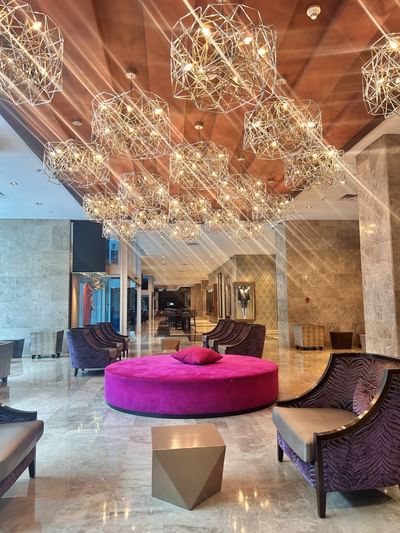 Round pink velvet ottoman set among purple armchairs under geometric pendant lights in the lobby at Megapolis Hotel Panama