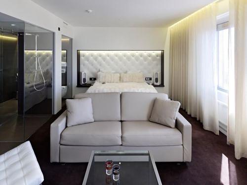 A modern hotel room with a taupe couch, glass table, and bed in a room with white walls, curtains, and dark floor.