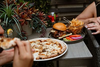 Table filled with delicious food including artisan pizza, bacon burger, french fries, and a cocktail served at Gattara
