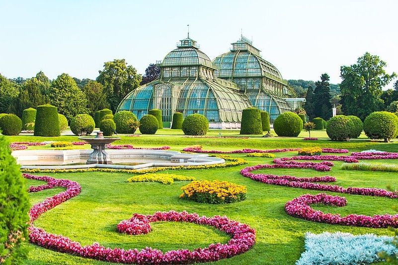 Palm House in the Schönbrunn Palace gardens with ornamental flower beds and manicured park landscaping on a sunny day in Vienna.