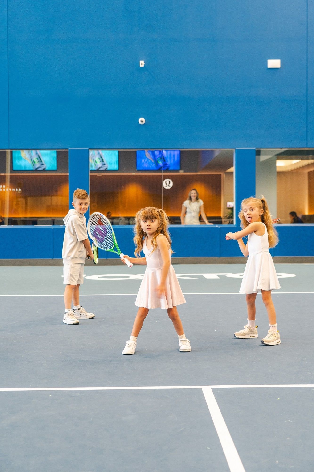 Three kids holding tennis rackets stand on an indoor blue tennis court with a large blue wall at Hotel X Toronto