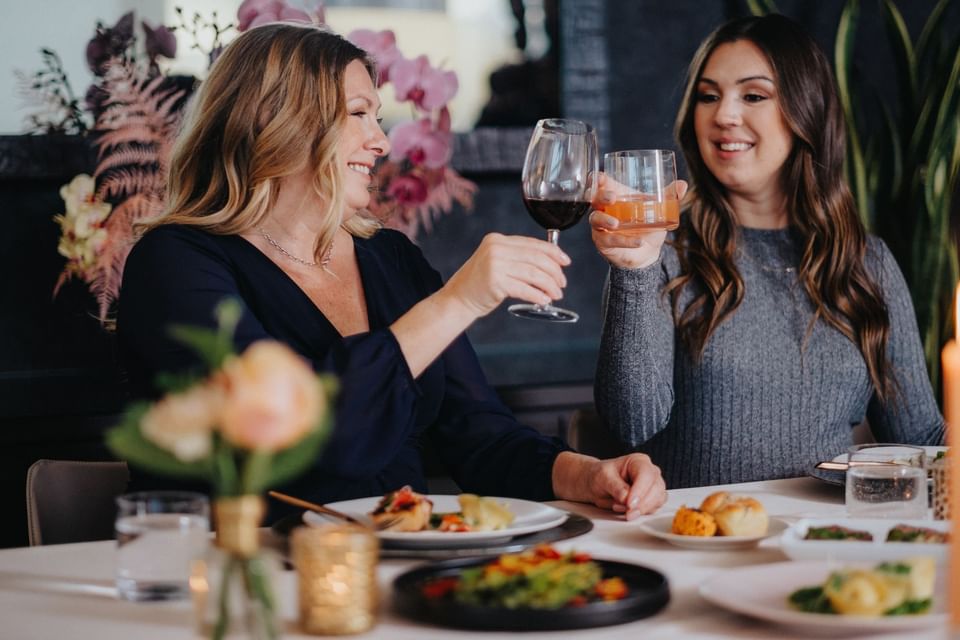 Two women raising glasses in a toast at a decorated dining table with gourmet food plates and flowers at Hotel X Toronto