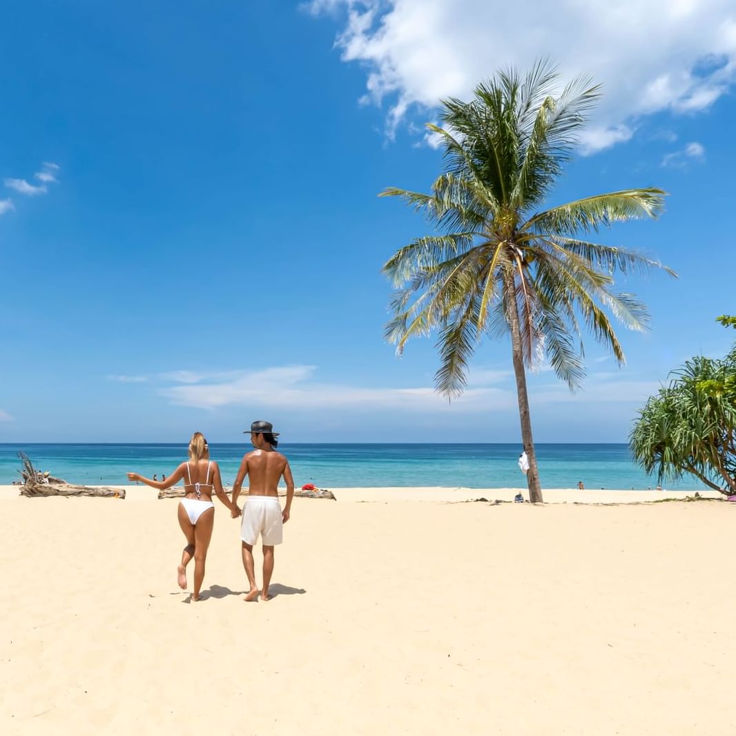 A couple is walking on the white sand Karon Beach on the sunny day with coconut tree on the right side