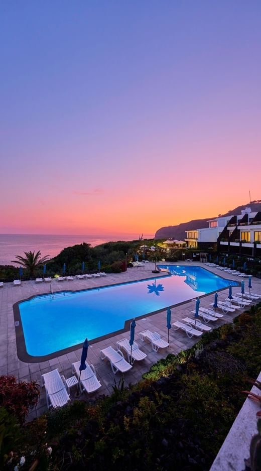 Illuminated outdoor pool and lounge chairs at dusk facing the ocean and hotel building at Caloura Hotel Resort