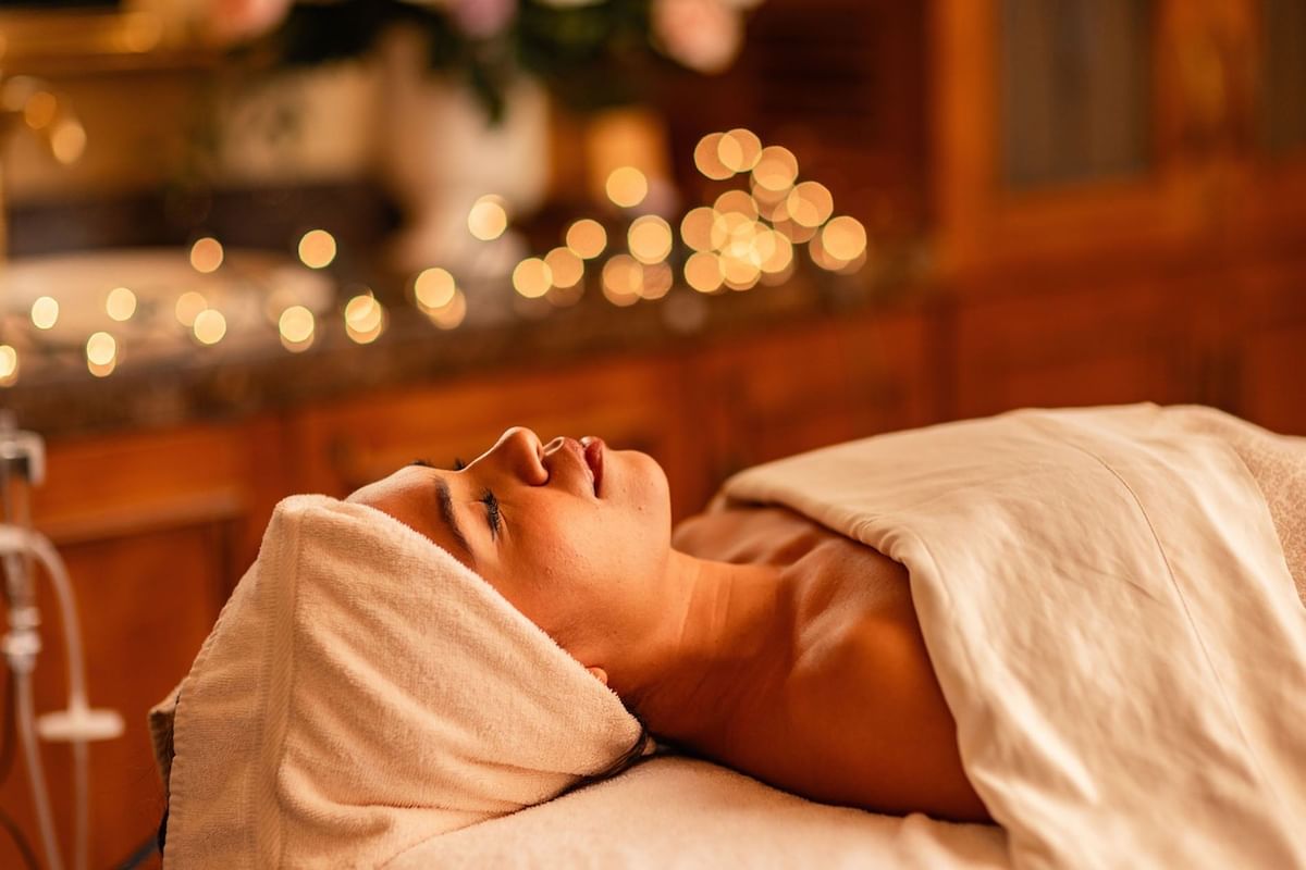 Relaxed woman receiving Fountain of Youth Signature Treatment with soft bokeh background lights and towels at Kelly's Spa