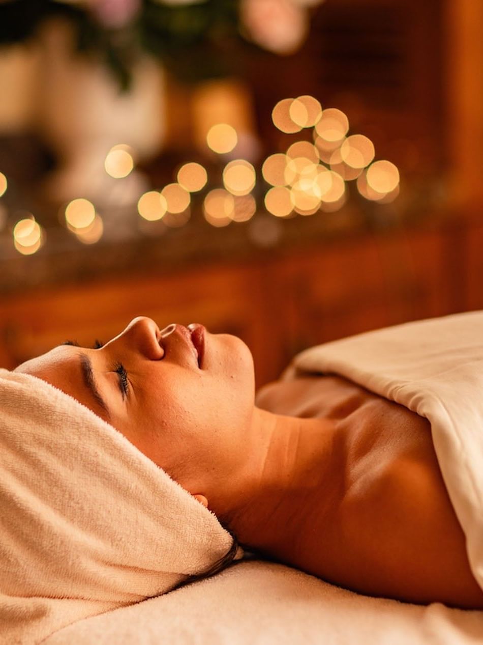 Relaxed woman receiving Fountain of Youth Signature Treatment with soft bokeh background lights and towels at Kelly's Spa