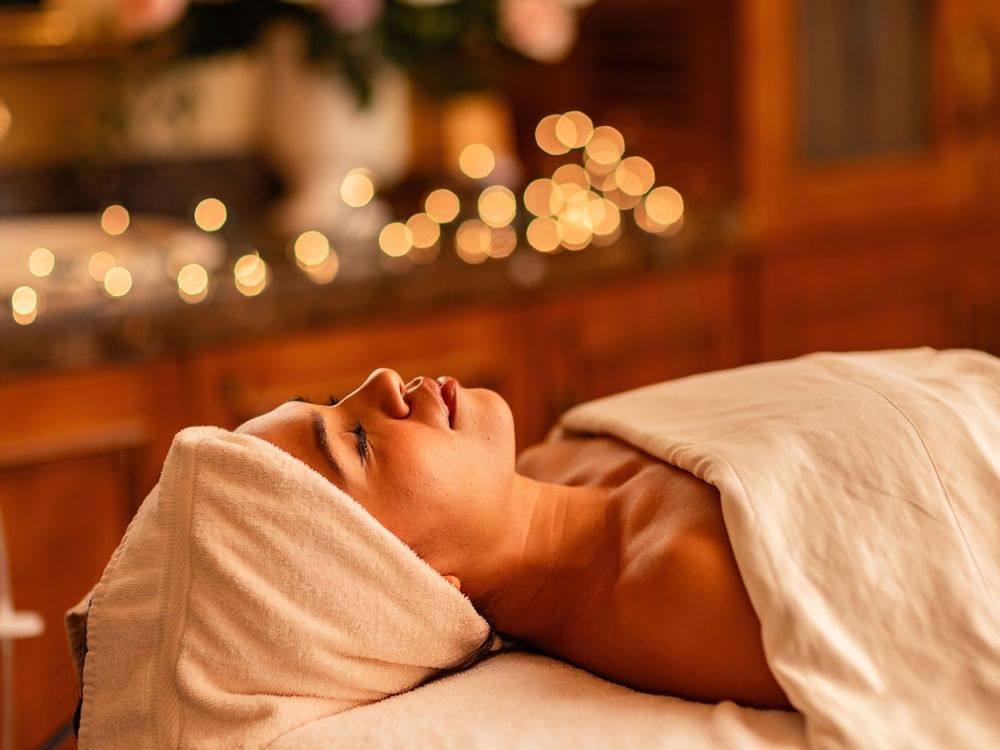 Relaxed woman receiving Fountain of Youth Signature Treatment with soft bokeh background lights and towels at Kelly's Spa