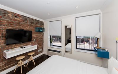 East Village Hotel room with a brick wall, flat screen TV, desk, stool, and bed with windows and a city view.
