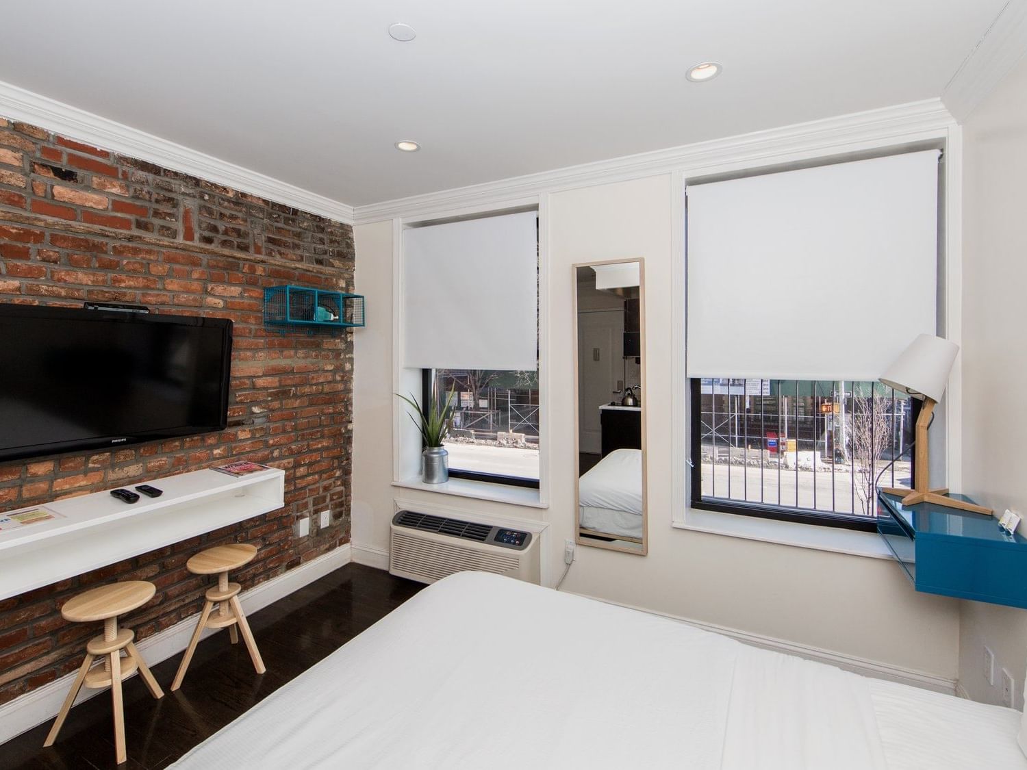 East Village Hotel NYC Queen Studio with a bed, brick wall, TV, stools, mirror, windows, and a city view.