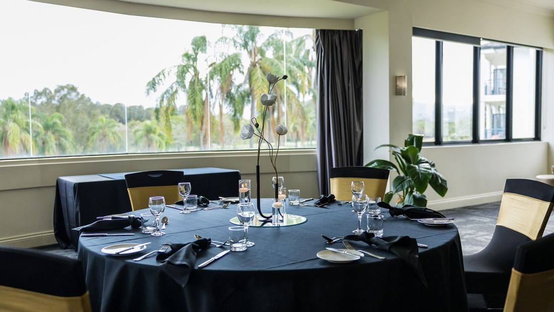 A dining room with round tables, black cloth, wine glasses, and chairs, with large windows and palm trees outside.
