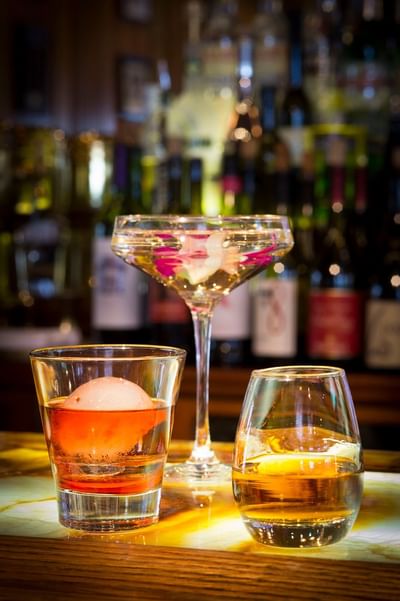 Three cocktails on a bar counter in Cascades Restaurant at The Stanley Hotel, with bottles of liquor in the background