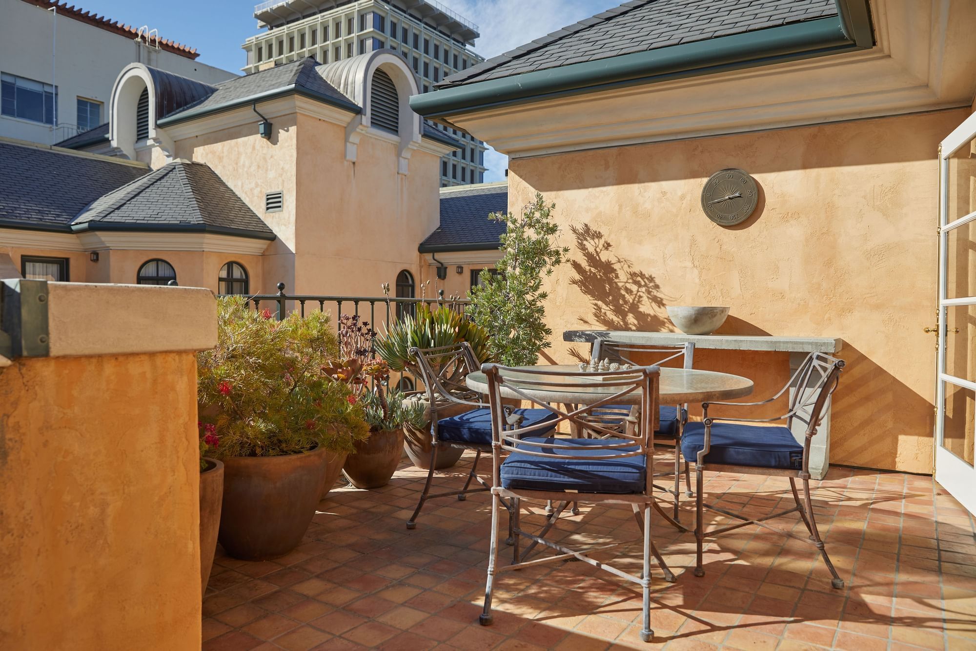 Sunny outdoor patio with metal chairs and potted plants overlooking buildings at El Prado Hotel
