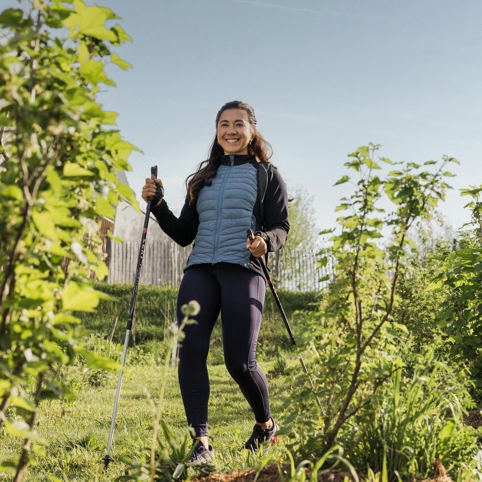 Eine Frau mit Stöcken lächelnd auf einem grünen Feld mit Pflanzen und einem Zaun im Hintergrund.