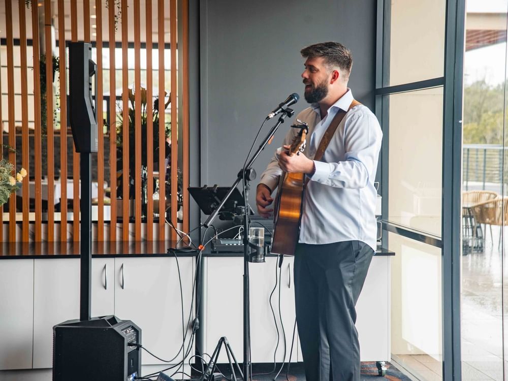 A man stands holding a guitar, singing into a microphone in an indoor setting with speakers and flowers.