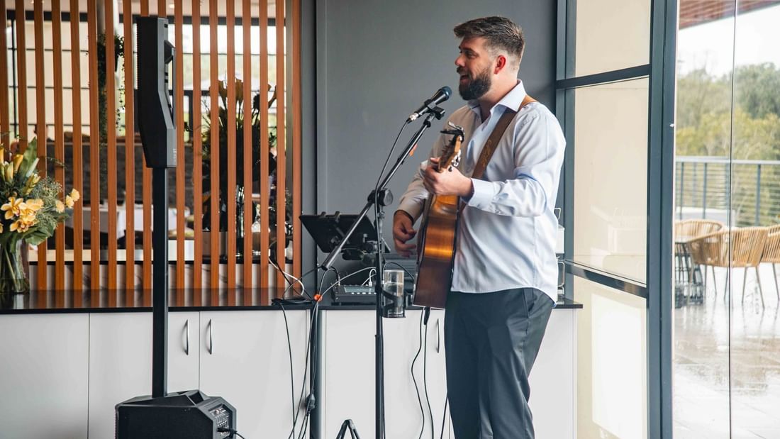 A man stands holding a guitar, singing into a microphone in an indoor setting with speakers and flowers.