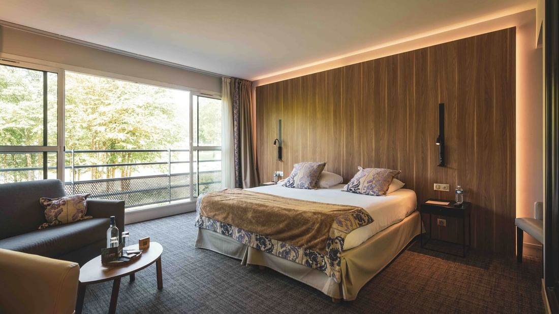 Chambre élégante avec lit double, canapé, table basse, et vue sur la forêt par une grande baie vitrée.