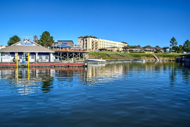 A lakeside view of a ship store, boat docks, and a large resort hotel under a blue sky at Shangri-La Resort and Golf Club
