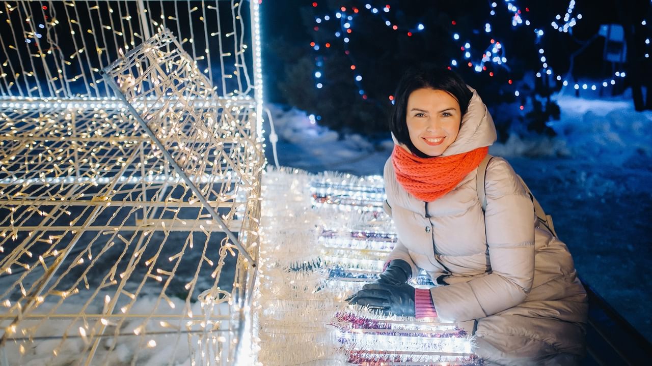 Woman in orange scarf smiling beside illuminated light display at Lions Festival of Lights during evening hours.
