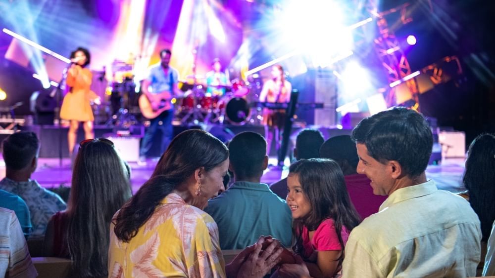 A family smiles at one another facing a colorfully lit stage where a band is performing.
