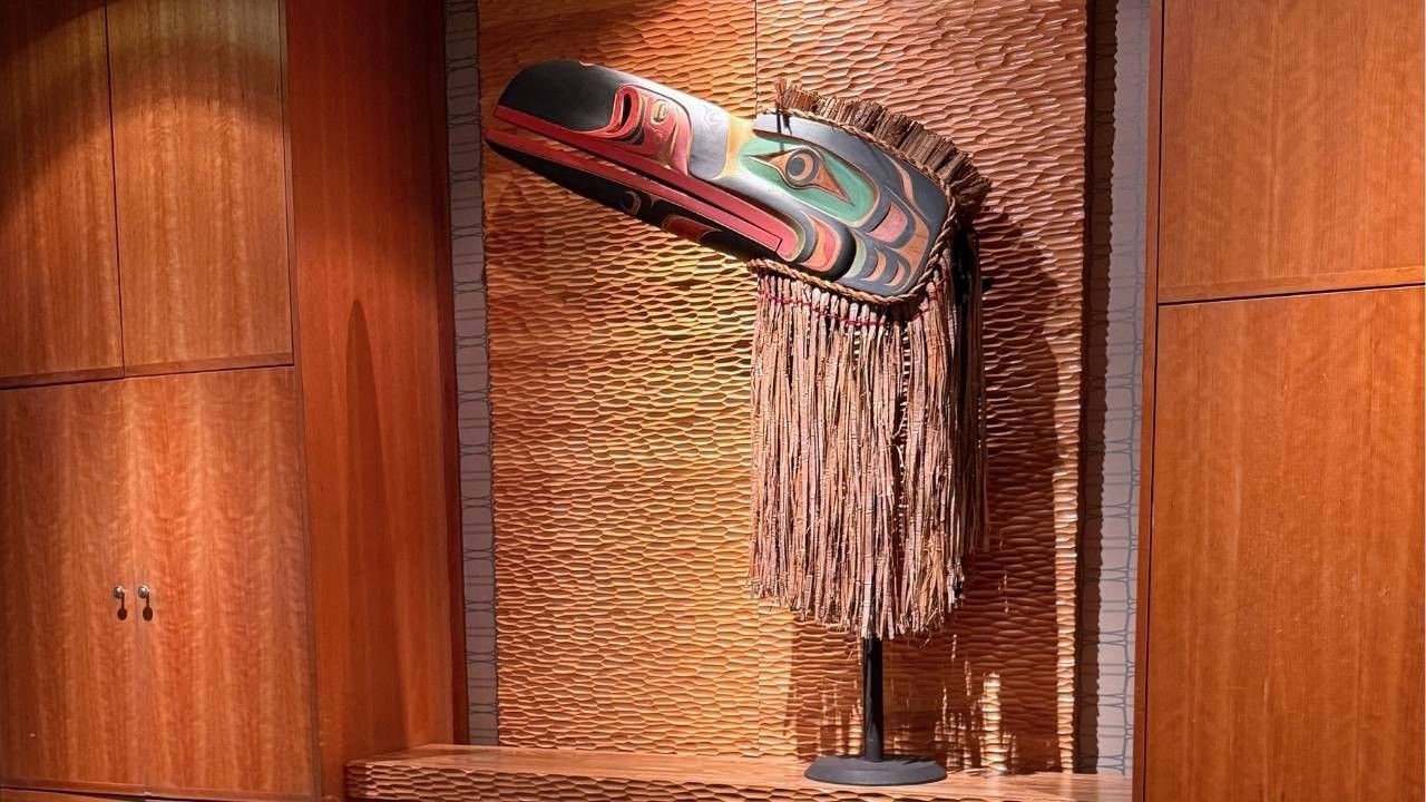 Indigenous raven mask with woven fringe displayed on a textured wood wall.