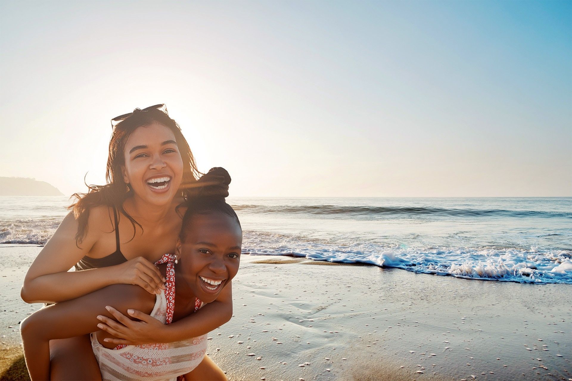 A happy woman smiles while hugging a young girl on a sunny beach, with gentle ocean waves washing onto the shore behind them.