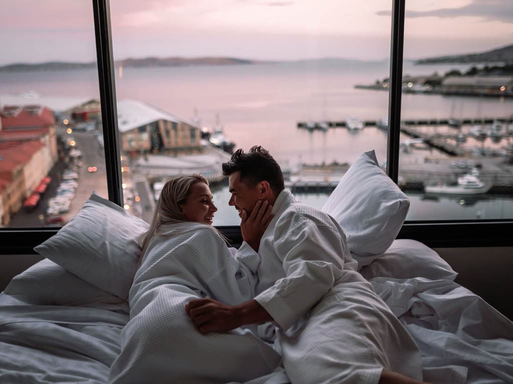 Couple wearing white bathrobes cuddling on a bed overlooking the bay at Hotel Grand Chancellor Hobart