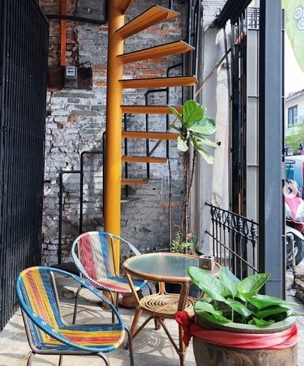 Colorful chairs and a table are arranged beside a spiral staircase against a brick wall at Kaya-Kaya Cafe near YTL Hotels