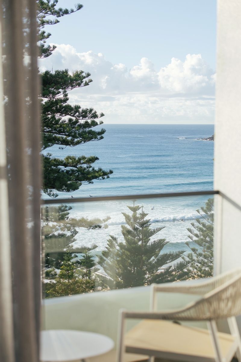 Elysium Manly balcony view revealing rolling ocean waves and pine trees framed by a sheer curtain next to a lounge chair