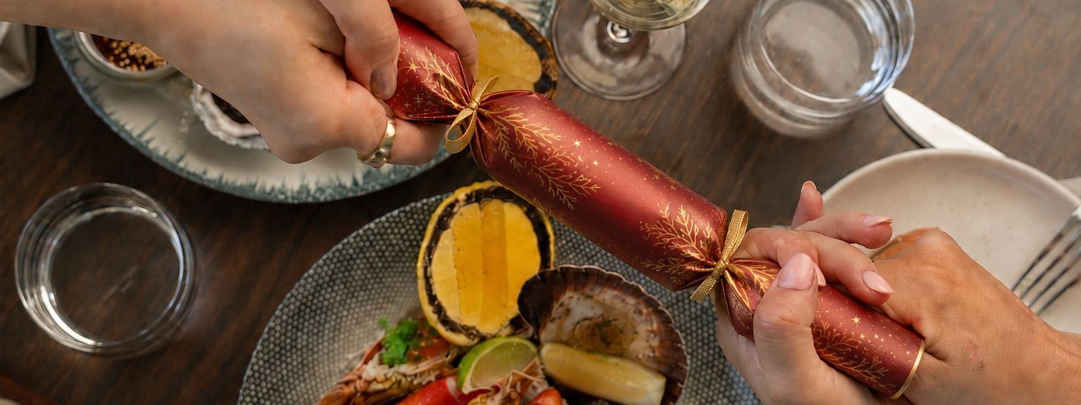 Two people pulling a festive red Christmas cracker over a table set with seafood, white wine, and holiday decorations.