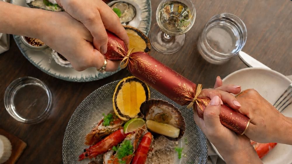 Two people pulling a festive red Christmas cracker over a table set with seafood, white wine, and holiday decorations.
