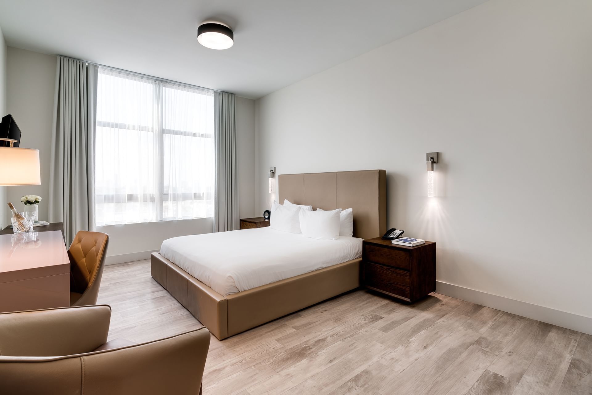 A modern hotel room with a tan leather bed frame, white bedding, and a large window.