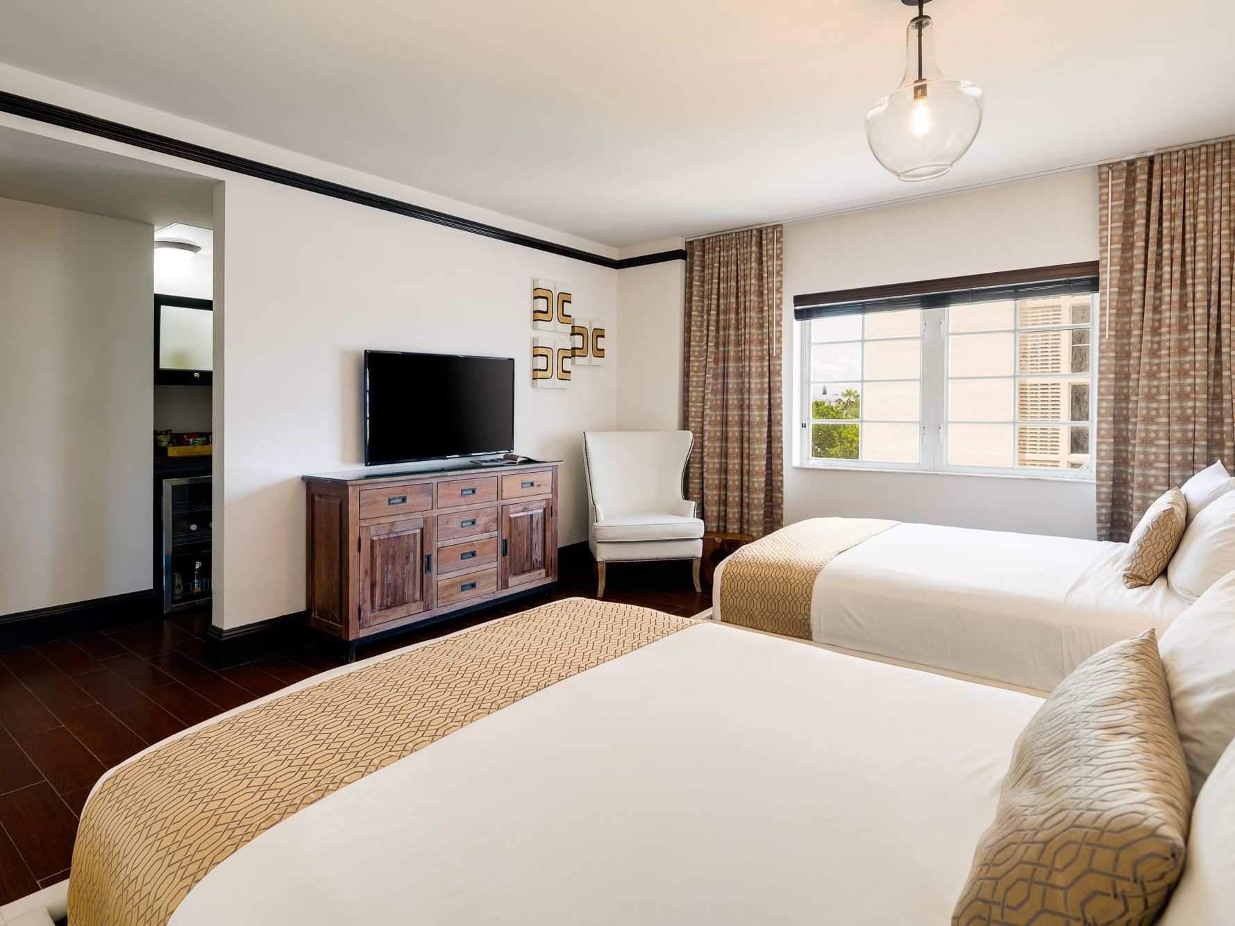 Two beds, wooden floor, TV on dresser, chair, curtains, lamp, and open door leading to another room.