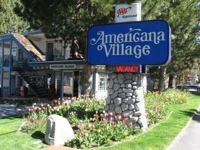 The blue Americana Village Suites sits on a stone base, surrounded by spring flowers, and the hotel's entrance on a sunny day