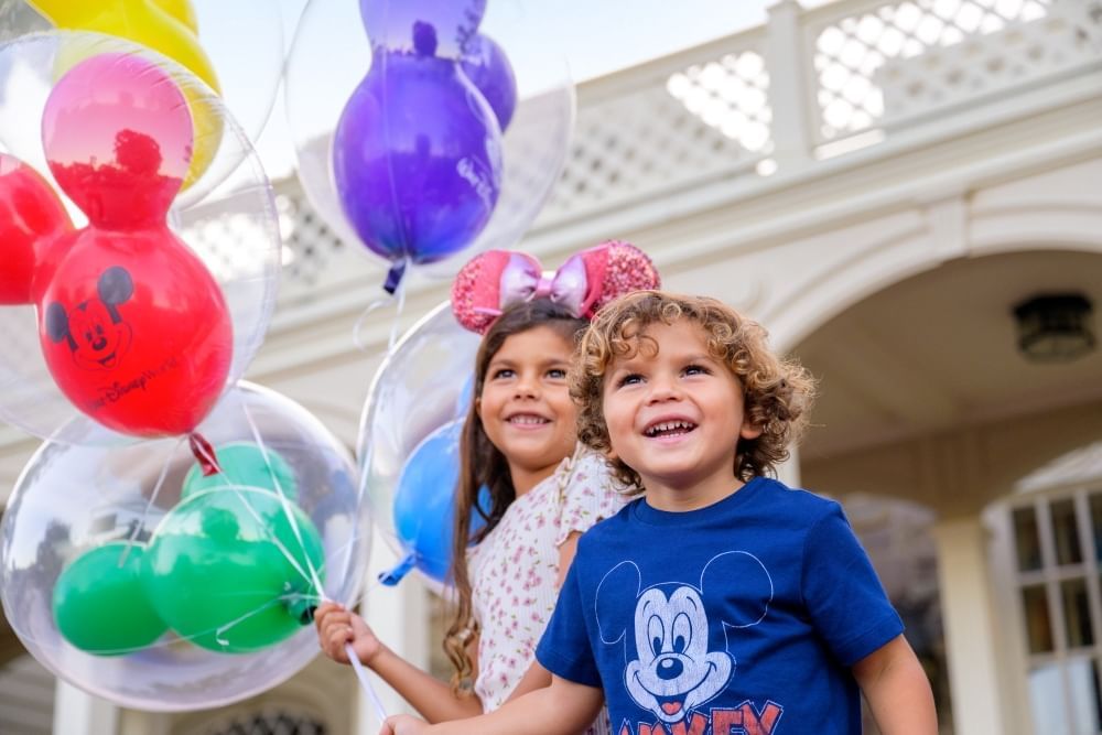 A boy and a girl smile outdoors while holding colorful Mickey balloons.