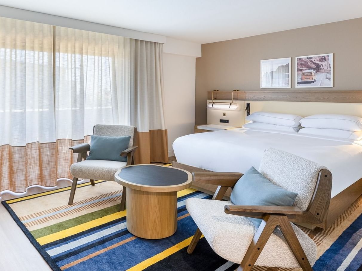 Modern hotel room with king bed, light wood furniture, blue accent chairs, and colorful striped rug
