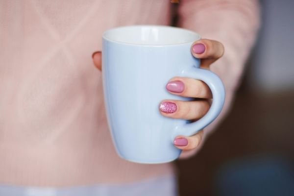 A close-up of a person wearing a pink sweater's hand with pink painted fingernails, holding a white mug. 